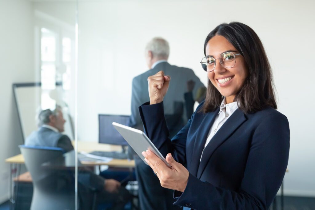 happy female professional in glasses and suit holding tablet and making winner gesture while two businessmen working behind glass wall copy space communication concept